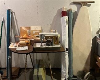 Lower Level:  A SINGER Touch 'n Sew sewing machine Model 603E includes its manuals and attachments.  The sewing table is separately priced.  On the left is a vintage movie screen; on the right are bolts of fabric and an ironing board. A vintage galvanized two-section bucket is also shown. 