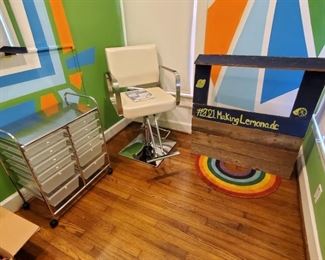White leatherette salon chair with metal base and adjustable foot pump and wooden lemonade stand with rainbow outdoor mat