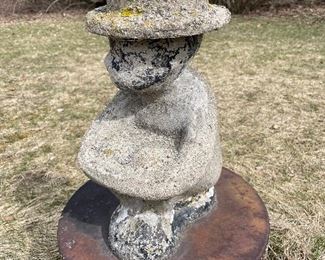 Cement figure of person sitting with removable cement hat. Some of the original black paint still on head and lower region of feet and butt.