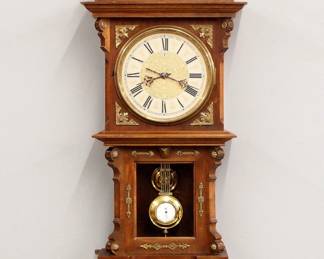 Lot 12: A turn of the century Junghans wall clock with unusual pull string winding.  8-day spring driven time and strike movement with engraved Silvered chapter dial, Arabic numerals and gridiron pendulum with porcelain indicator.  Walnut case with arched crest and carved finials, circular Brass dial door and glass lower with fluted pilasters and shaped bracket drop.  Old finish with some wear, flake in dial glass, running when cataloged.  40 1/2" high overall.  ESTIMATE $800-1,200
