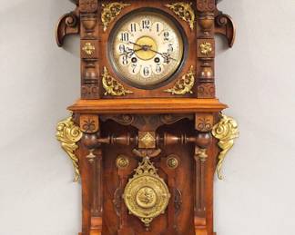 Lot 24: A turn of the century Lenzkirch open well wall clock.  Series 26, 8-day spring driven time and strike movement with embossed Brass dial, Silvered chapter ring with Roman numerals and fancy Brass pendulum, serial #1 Million 111254 indicates a production date of 1896.  Walnut case with applied Cast Brass decoration and carved detail, arched crest over molded crown and dial door with circular glass, open well with turned rail over a lower bracket drop.  Older refinishing with minor wear, lacks one small Brass detail, running when cataloged.  37" high overall.  ESTIMATE $800-1,200
