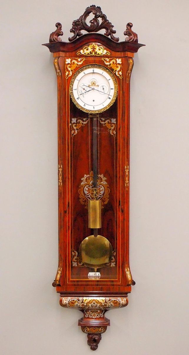 Lot 68:  A Very Good quality Inlaid Vienna Regulator wall clock by "J. Thiel, in Wien".  60-day weight driven time only movement with a flat porcelain dial, with visible escapement, cut Steel verge and subsidiary Day/Date dials, Roman numerals and Cast Piecrust bezel, marked "J. Thiel, in Wien".  Transitional Style Rosewood case with Brass, Copper and Pewter marquetry inlay features a carved crest above an arched carved crown with inlaid frieze, an arched door with chamfered corners and fluted columns with inlaid medallions, inlaid back panel and shaped inlaid drop and carved lower finial.  Finish in excellent condition with minor loose inlays, running when cataloged.  54 1/2" high overall.  ESTIMATE $4,000-6,000
