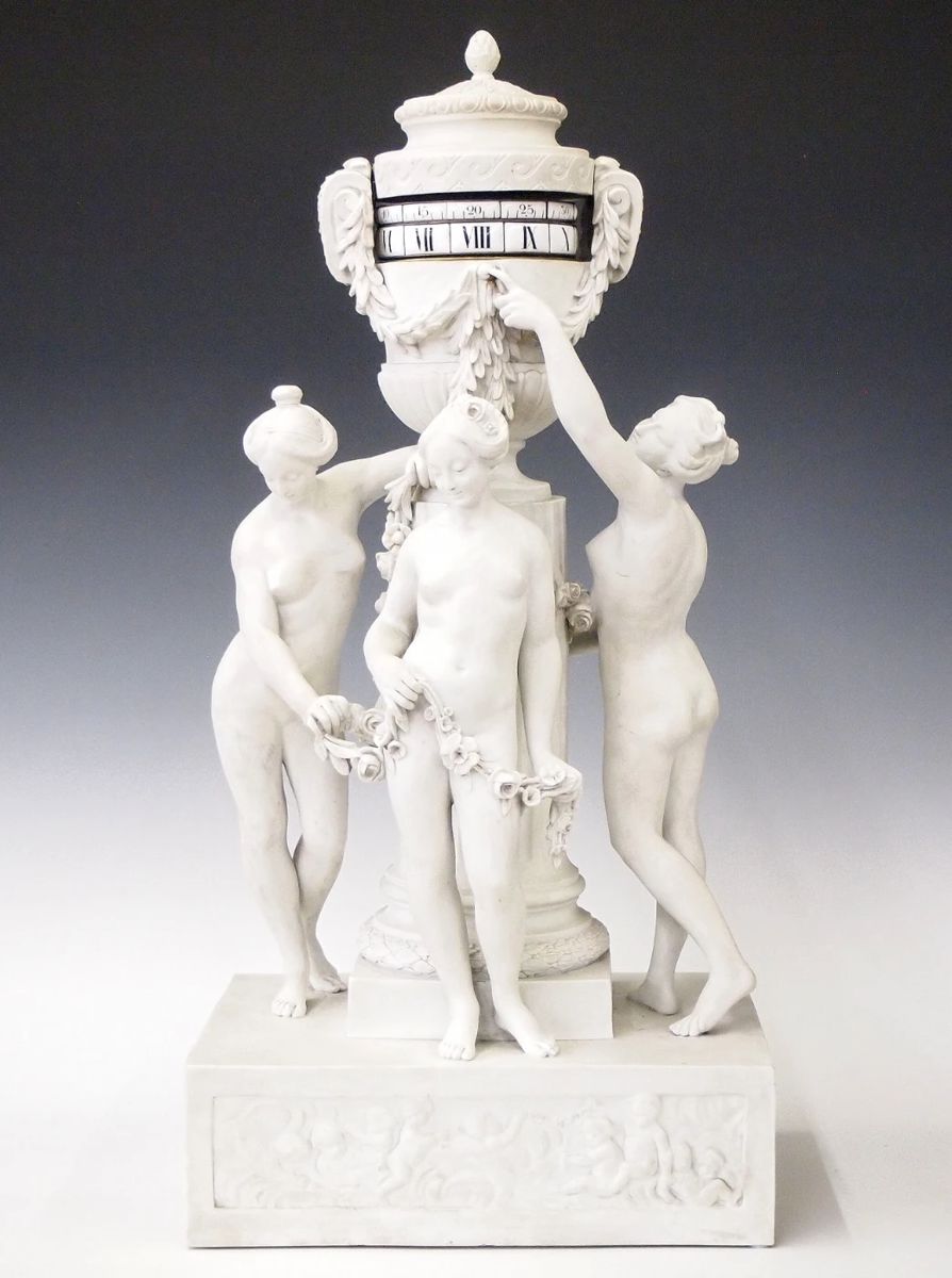 Lot 72: A late 19th century French Three Graces Annular clock.  8-day time and strike movement with platform escapement and dual annular dial rings with porcelain markers, Roman hour numerals and Arabic minutes.  Bisque porcelain case with upper urn and lift off cover and foliate detail, over Neoclassical figures of the Three Graces with floral swags standing before a fluted pedestal on a rectangular plinth with bas relief panels of frolicking Cherubs.  Minor wear, no damage noted, running when cataloged.  24 1/2" high overall.  ESTIMATE S4,000-6,000
