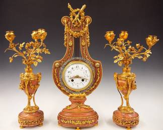 Lot 71: A late 19th century French three piece Gilded Bronze and Marble Lyre clock set.  8-day time and strike movement with knife edge suspension, convex porcelain dial, Arabic numerals with hand painted floral swags and Gilded filigree hands, fitted with a gridiron pendulum and swinging exterior bezel decorated with applied "brilliants", serial #217.  Louis XVI Style Rose Marble case with Gilded Bronze frame with swags and foliate detail, surmounted by torches, on a foliate pedestal base with a pair of three branch candelabra with foliate arms, and tripod bases with Ram's head and foliated swag detail.  Minor wear, hairline free dial, minor fissures in stone, movement complete and running when cataloged, set-up and balancing will be required after moving.  18 3/4" high overall.  ESTIMATE $4,000-6,000
