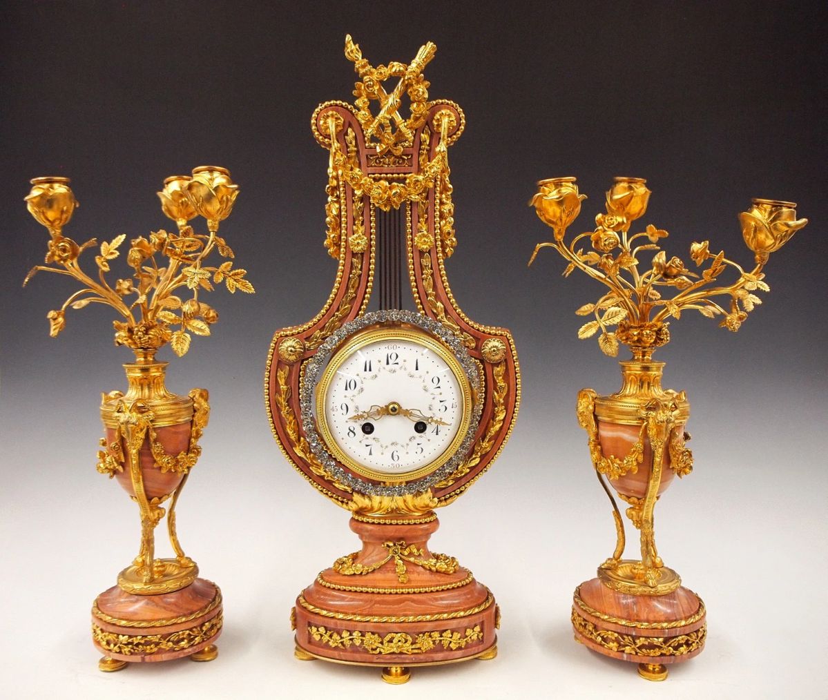 Lot 71: A late 19th century French three piece Gilded Bronze and Marble Lyre clock set.  8-day time and strike movement with knife edge suspension, convex porcelain dial, Arabic numerals with hand painted floral swags and Gilded filigree hands, fitted with a gridiron pendulum and swinging exterior bezel decorated with applied "brilliants", serial #217.  Louis XVI Style Rose Marble case with Gilded Bronze frame with swags and foliate detail, surmounted by torches, on a foliate pedestal base with a pair of three branch candelabra with foliate arms, and tripod bases with Ram's head and foliated swag detail.  Minor wear, hairline free dial, minor fissures in stone, movement complete and running when cataloged, set-up and balancing will be required after moving.  18 3/4" high overall.  ESTIMATE $4,000-6,000
