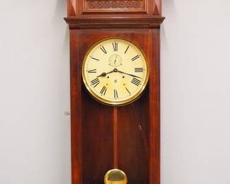 Lot 115: A late 19th century Waterbury "No. 54" type Regulator wall clock.  8-day dual wind spring driven time only movement with a painted metal dial, Roman numerals and subsidiary seconds.  Walnut case with a shaped crest and turned finials above a paneled frieze with carved detail and a single long molded door with arched glass over a carved bracket drop.  Older refinishing with minor wear, restored dial, running when cataloged.  58" high overall.  ESTIMATE $800-1,200

