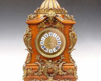 Lot 135: A late 19th century Lenzkirch Louis XV Style table clock.  Series 52, 8-day spring driven time and strike movement with cast Brass dial and Blue Roman numerals on porcelain markers, filigree hands and cast Brass bezel, serial #816856 indicates a production date of 1888.  Louis XV Style Bird's Eye Maple case with domed top and extensive cast Brass appliques which continue onto the sides of the shaped case, includes a woman and Cherub on Lion's mask feet.  Old finish with slight wear, shrinkage cracks, tiny flakes on 8:00 marker, running when cataloged.  20" high overall.  ESTIMATE $800-1,200
