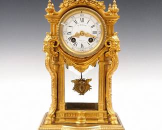 Lot 138:  A turn of the century French mantel clock by "A. D. Mougin, Paris" retailed by "P. Hubeau, Namur (Belgium)".  8-day time and strike movement with convex porcelain dial, Roman hours and Arabic minute with fancy Gilded hands.  Gold finished Spelter case with shaped top, Ram's Head columns and beveled glass on a White marble base with beaded feet.  Some wear, flakes at winding arbors, running when cataloged.  15" high overall.  ESTIMATE $600-800
