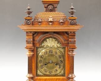 Lot 144:  A late 19th century German Bracket clock.  8-day spring driven time and strike movement with engraved, arched Brass dial and Arabic numerals, cast Brass applique and spandrels.  Walnut case with turned finials on a shingled "roof" with Lion's mask detail, over a single door with arched top and turned fluted columns and carved lower frieze and a molded base.  Older finish with minor wear, running when cataloged.  27 1/4" high overall.  ESTIMATE $600-800
