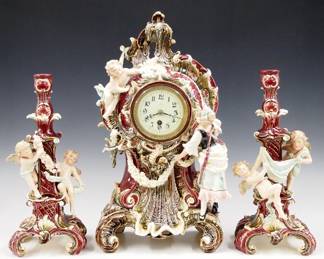 Lot 145: A turn of the century Junghans three piece clock set.  8-day time only movement with lever escapement and flat porcelain dial with Arabic numerals and Crossed Arrow Trademark.  Louis XV Style Earthenware case and candelabras, features a clock with Cherub and Maiden on a scrolled base with floral swags, and a pair of candelabras with Cherubs, all with multi-color glaze decoration and Gilded detail.  Some wear, Cherub on the clock lacks a wing and one on a candelabra has a replaced wing, candelabras lack upper branches, clock running when cataloged.  Up to 19 1/2" high overall.  ESTIMATE $400-600
