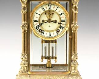 Lot 189: A late 19th century Ansonia "Martha" Model Crystal regulator.  8-day spring driven time and strike movement with visible escapement and two part porcelain dial with Roman numerals, cast Gilded bezel and faux Mercury pendulum.  Fancy cast Brass case with scrollwork panel on dentil molded top, beveled glass doors and sides flanked by fluted columns on a stepped base with scrolled feet.  Worn Nickeled finish, running when cataloged.  11 1/2" high overall.  ESTIMATE $400-600

