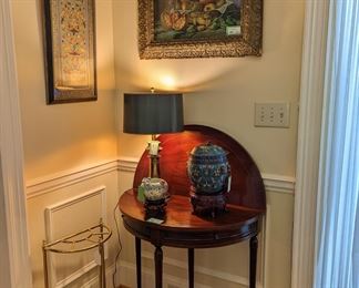 Vintage mahogany Hickory Chair demilune game table, amazing Chinese cloisonne lidded urn on wooden stand, Asian porcelain bowl on wooden stand, vintage Stiffel brass table lamp, antique fruit still life in gold wooden frame, one of a pair of framed Asian silks and vintage brass/cast iron umbrella stand.