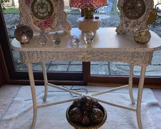 White wicker table, vintage clocks and oil lamp with spotted cranberry shade