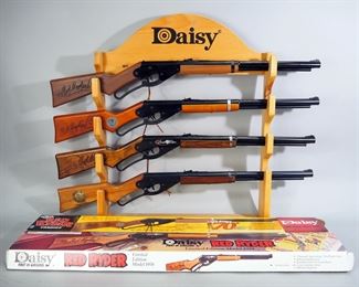 Daisy Red Ryder Model 1938B Anniversary Edition BB Guns, Qty 4, Includes 1938-1988 Edition, 60th Anniversary (With Box), 65th Anniversary, And 70th Anniversary (With Box), On Daisy Gun Display Rack