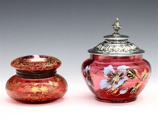 Two turn of the century Cranberry glass covered jars.  Enameled floral and figural decoration with Gilded detail, one with cast metal cover and one with hinged top.  Some surface wear, minor losses to decoration.  Up to 6" high.