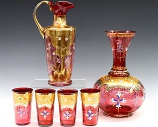Six pieces of late 19th century Cranberry glass drinkware.  Enameled floral decoration with Gilded detail.  Includes a pitcher with ruffled rim and a carafe with four matching glasses.  Some surface wear and minor losses to Gilding, a few tiny flakes at rims, carafe with production flaw at neck.  Up to 9 1/4" high.