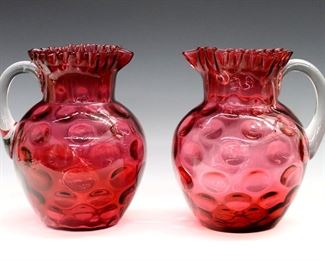 Two turn of the century Cranberry glass pitchers.  Bulbous forms with applied clear handles, ruffled rims and thumbprint decoration.  Some surface wear, one with production imperfections at body.  Each 8 1/2" high.