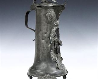 A late 19th century German Pewter Flagon attributed to August Weygang.  Hinged cover with rampant lion shield and a shaped thumb piece with scrolled handle, over a coat of arms and inset musician figure on a molded base with three cherub mask legs.  Marked "A W" with "1870" inside legs.  Some damage and distortion at lid, minor denting.  14" high.  