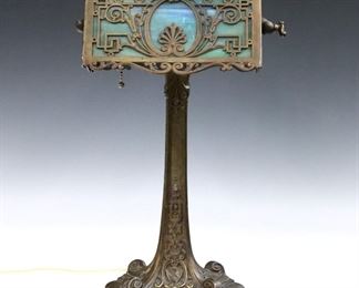 A turn of the century American Arts & Crafts Bronze desk lamp by Wilkinson, New York.  Adjustable cast filigree shade with Blue/Green Slag Glass panel on a stylized cast base with Gilded finish.  Marked "Wilkinson, Brooklyn, NY" at underside. 