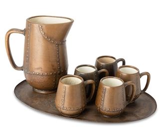 1042
Circa 1910s
A Clewell Copper-Clad Ceramic Cider Set
Pitcher and mugs each marked to the underside: Clewell / Canton, Ohio
The set comprising one handled pitcher (10.25" H) and five mugs (each: 4.5" H), each ceramic with faux-riveted copper overlay, all set upon a solid copper oval tray (19.25" Dia.), 7 pieces
Estimate: $300 - $500