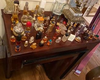 Probably at least 75 small collectible perfume bottles on top of an vintage sewing machine in an art-deco influence designed desk with matching bench seat