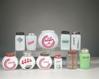 (11PC) GLASS SUGAR & SPICE SHAKERS | Including various circular milk glass sugar shakers with red paint, cinnamon shaker with image of a dog, square pepper & flour shakers and more. l. 4 x w. 5.5 x h. 5 in