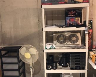 Lower Level-Storage Room:  A short plastic rolling drawer unit is to the left of a floor model fan and a tube shelf unit.  On the shelf are:  a portable workbench, metal cube storage, window fan, two motherboards, and more.