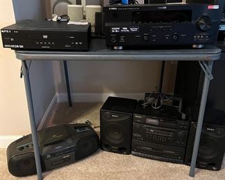 Lower Level:  On top of the table are an APEX DVD player; one of two YAMAHA R-V765 AV Receivers; and several small speakers.  Below are a SONY CFD560 radio-CD-cassette recorder and a SONY "boom box." 