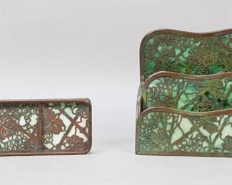        334	Tiffany Studios Grapevine Pen Tray & Letter Rack	2 Signed Tiffany Studios Grapevine pattern desk pieces. Letter rack, 10"L x 2 1/2"D x 6 1/4"H; pen tray, 9 5/8"L. Patinated bronze with green slag glass. Both stamped Tiffany Studios New York. Letter rack glass cracked on back and middle sections, losses and scratches to bronze patina, back section bent and possibly repaired; pen tray losses and scratches to bronze patina.
