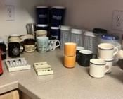 A wide assortment of mugs, individual and set, and large water cups with straws. The red container in the front is a canister for small cans of cleansing powder with a cameo accent.