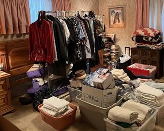 In the back of this picture is the rack of mostly men's clothing, a headboard, a couple of nightstands, and lots of bedding.