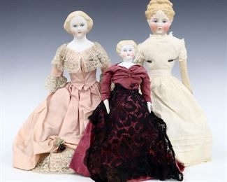 Three 19th to early 20th century dolls. Bisque heads with Blonde molded hair and painted detail on cloth jointed bodies. Includes a doll with porcelain lower arms and legs wearing a Pink dress with lace, a doll with pierced ears wearing a White dress with lace, and a smaller doll with porcelain lower arms and legs wearing a Mauve dress with lace. Some wear to paint, fabric with some fading, staining and small holes, doll in white dress with repaired head. Up to 19" long. ESTIMATE $100-200