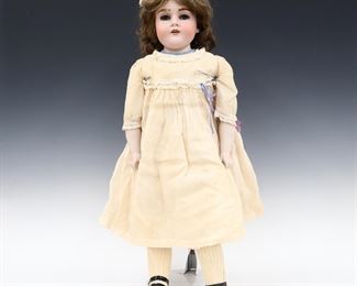 A turn of the century JD Kestner doll. Full bisque head with real Light Brown hair, sleep eyes with hair eyebrows and eyelashes, open mouth with teeth and painted detail. On a kid jointed body with porcelain lower arms, wearing a White Swiss point dress with lace and patent leather shoes. Original "JDK Germany" sticker at torso. Minor wear to paint, some losses to eyebrows, one leg losing stuffing, fabric with some fading and staining. 29" long. ESTIMATE $200-300