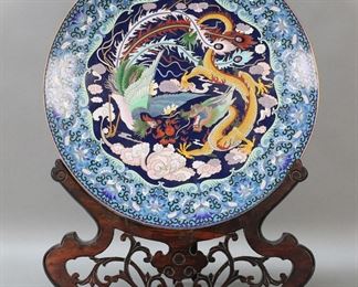 242	Chinese Cloisonne Charger on Stand	Chinese cloisonne charger, dragon and phoenix motif. On wooden stand. Charger 15 3/8"-diameter, with stand 20 1/4"H. Dent to rim of charger at top, minor losses to wooden stand.

