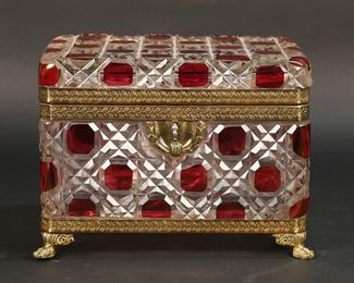 321	Cranberry Cut to Clear Dresser Box	Cranberry color to clear cut glass dresser box. Gilt bronze mounts, paw feet. Unsigned. 7 1/2"W x 5 1/2"D x 5 3/8"H. Flake just below hinge, flake to one side, internal chip to glass on underside, glue residue present around mount on underside of box.
