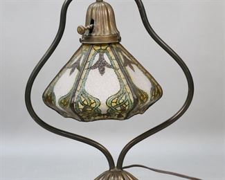 393	Desk Lamp With Reverse Painted Shade	Arts and Crafts style desk lamp. Reverse painted chipped ice shade. Bronze harp form base. Shade and base appear unmarked. In working order. Some damage to metal socket cover where switch goes in. Switch is loose. Nicks and wear to lamp base. One panel of shade has dark staining on bottom. 15" Tall.
