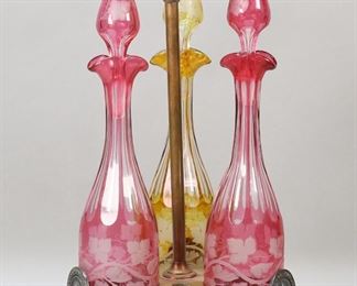 398	Bohemian Glass Triple Cruet Set	Boehmian glass cruet / decanter set. Three color to clear cruets, two cranberry and one amber, all with grape and leaf motif. In a brass holder with Greek key motif and Greek medallions. Each cruet 15 7/8"H, holder 19 3/4"H x 9 3/8"W. Discoloration and losses to brass patina throughout, scratches to the undersides of all three decanters, tips of stoppers on both cranberry cruets possibly ground down, interior of one cranberry stopper cloudy.
