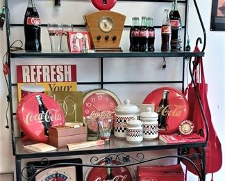 Bakers rack just full of Coca Cola collectables. See the REFRESH sign? On both ends of that shelf those round things are telephones, and there is a new one in the box