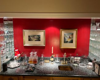 Family Room-Wet Bar:  Two M. Charles Rhinehart original pieces of art hang on the wall.  They are flanked by glass shelves of crystal (Baccarat, Riedel, Waterford, Tiffany & Co., and others).  A four decanter Tantalus and a glass V.P. 1985 champagne bucket are among other barware on the counter.