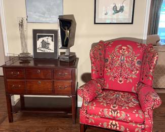 Family Room:  A petite sideboard with a brass galley has two drawers.  It is to the left of a raspberry/bird theme embroidered wingback chair.  