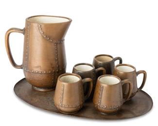1015
Circa 1910s
A Clewell Copper-Clad Ceramic Cider Set
Pitcher and mugs each marked to the underside: Clewell / Canton, Ohio
The set comprising one handled pitcher (10.25" H) and five mugs (each: 4.5" H), each ceramic with faux-riveted copper overlay, all set upon a solid copper oval tray (19.25" Dia.), 7 pieces
Estimate: $200 - $400