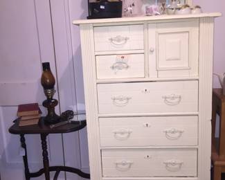 An antique chest of drawers with a small door to conceal hats. 
The half-round table, left, is topped with a lamp, vintage books and a mirror. 