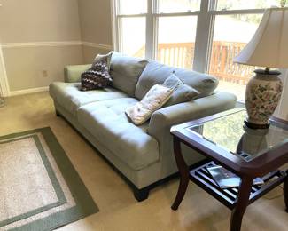Modern light gray sofa, green and beige area rug, floral ceramic lamp, wood and glass top end table, various throw pillows