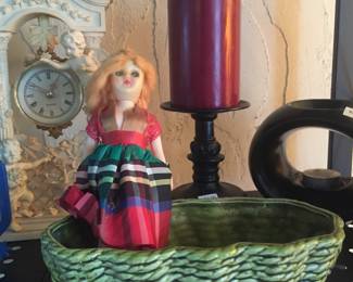 A jointed doll with moveable eyes sports a plaid taffeta dress while nestled in a vintage planter. A resin clock and candle holders round out the scene.