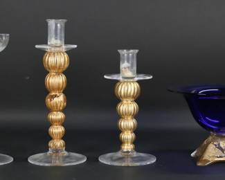 132	4 Pieces Art Glass Willsea O'Brien, Rose Tattoo	4 pieces Murano style art glass. Willsea O'Brien pedestal bowl, cobalt, with gilt decorated base, signed on the underside, 6"H x 8 7/8"-diameter; 2 Rose Tattoo candlesticks, Venetian style with gilt decorated stems, each signed on the underside, taller 12'H; unsigned Venetian style candlestick with gilt decorated twist stem, 10 5/8"H. Bowl repaired at center, scratches to feet, flake to one foot, chips to two feet; losses to gilt decoration on base of bowl and stems of all three candlesticks, candlesticks all with wax residue.
