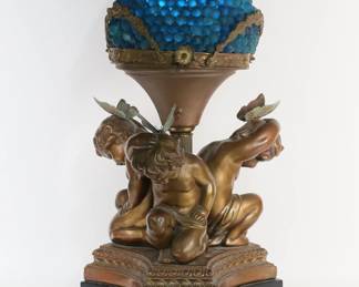 446	Armor Bronze Clad Cupid Lamp Glass Grapes Shade	National Metalizing Company "Armor Bronze" bronze clad figural Cupid lamp. With a blue glass grape shade. Later butterflies added to Cupids' heads. Mounted on a wooden base. Armor Bronze and copyright marks on the bronze clad base. 30"H including shade, 15"W. Fitting for shade bent and with cracks to rim, shade does not sit flush on base, crack to top of one Cupid's head, chips to corners of bronze clad base, loss to bronze patina throughout; paint loss, chips and scratches to wooden base.
