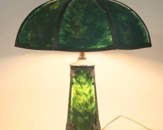 422	Reverse Painted 6 Panel Lamp	6 panel reverse painted table lamp, with 3 panel reverse painted base with patinated metal overlay with floral motif. Textured glass on shade and base. Lamp and shade both unsigned. Reverse painting on base and shade both covered by later green painting. Lighting and lighting in base both work. 21 3/4"H to top of cap, shade 19 1/4"-diameter. Later green painting to interior and exterior of shade and interior of base, one glass panel on shade replaced with painted cardboard, minor chips to edges and corners of glass on interior of shade, fraying to cord, losses to patina on pipe.
