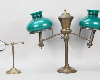 412	Brass Double Arm Student Lamp & Magnifier	Brass double arm electrified student lamp with emerald green cased glass shades, 19"H x 24"W. With a brass magnifier on stand, adjustable, 9 1/4"H. Lamp both arms repaired at lamp fittings and one loose, dents to cap and base, repairs at top, chips and fleabites to rims of both shades; screw missing to hold arm of magnifier in place.
