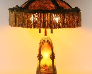 421	Reverse Painted Table Lamp Landscape Scene	Gilt metal reverse painted table lamp. 4 panel light-up base with reverse painted scene of houses in landscape, 5 panel shade with later painted plastic panels and silk fringe. Unsigned. 25 3/4"H including finial, shade 17 3/4"-diameter. Losses to gilt patina on base and shade, some paint loss to shade, no bottom on lamp base.
