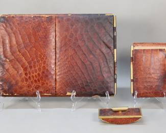 411	3 Piece Alligator Leather Desk Set	3 piece alligator leather desk set. Rocker blotter, note pad and blotter. Blotter 19 1/2"L x 13 1/2"W closed (open 38"L). Blotter and notepad tears and losses to leather along edges, at corners and at hinges, notepad cover leather straps missing at top and bottom of hinge and cover partially detached, staining to interior; blotter pen marks, fading and discoloration to interior of covers and surface of blotter, tear to leather on cover; rocker blotter some wear and discoloration to paper on base, dents to brass and losses to leather under brass rim.
